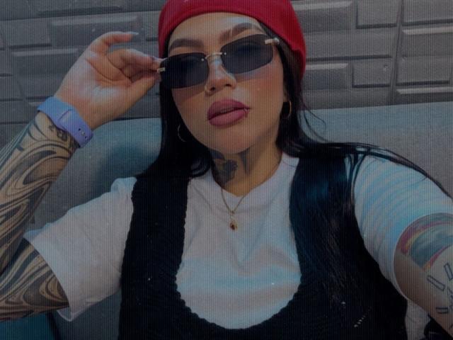TessaMagic with tattoos, red beanie, black sunglasses, white tee, black vest, sticking out tongue, gold necklace, blue wristband, grey background, holding sunglasses, looking at camera.