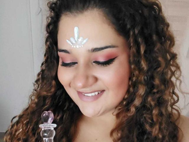 NataliaWendys has curly brown hair with blonde highlights, smiling down. She has pink eyeshadow, black eyeliner, and a white symbol on her forehead. Her eyes are closed, and she holds a pink glass object with a rounded top. Her skin is light brown, and her lips are pink. The background is out of focus. The image focuses on her face and the glass object. Slight shadow on her cheek adds depth.