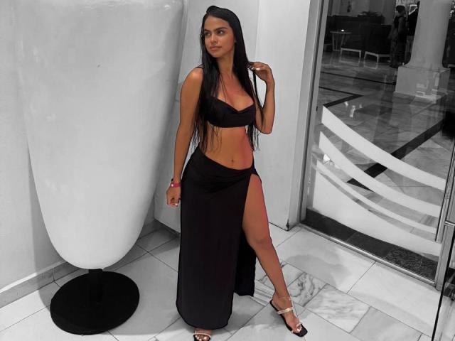 AlannaThompson has long black hair, a black bra, and a black skirt with a high slit, showing her bare ass. She's wearing silver heels, standing indoors.