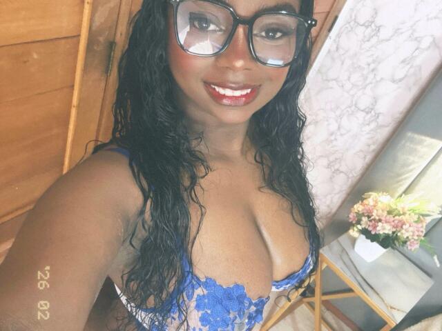 SamyLorenss, a dark-skinned woman with curly black hair, wears large black glasses and a blue lace bra that highlights her large breasts. She has a bright red lipstick and a broad smile, looking directly at the camera. The image is a selfie, with a wooden door and a floral arrangement on a small table in the background. The watermark "ZNG SSM" is in the lower left corner. The lighting is warm, emphasizing her skin tone and the texture of the lace.