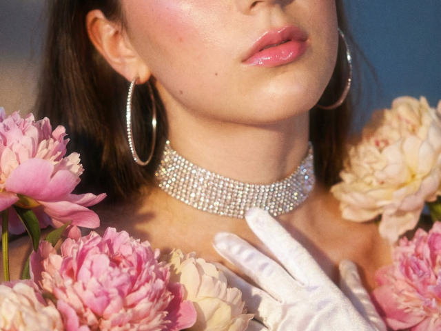Polinochka's glossy pink lips, sparkling choker, and white gloves are surrounded by pink and white peonies, with large silver hoop earrings. Her chin and neck are visible.