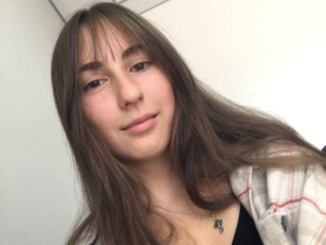 Polinochka has long brown hair, bangs, and a subtle smile with pink lips. She's wearing a plaid shirt over a black top, showing a small necklace.