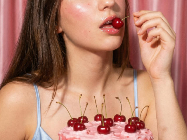 Polinochka with shiny pink lips and brown hair, holding a cherry near her mouth, wearing a blue spaghetti strap top, has cherry-topped pink cake in front of her.