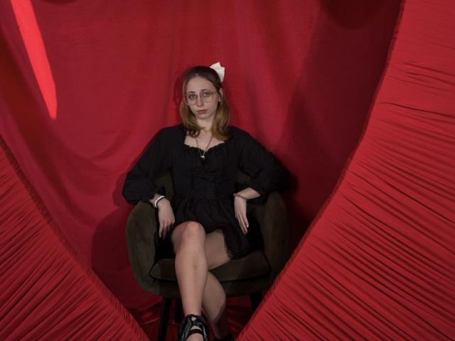 TracyLeex with glasses, black dress, and white bow, sitting in green chair, legs crossed, red draped background, black shoes, looking slightly annoyed, hands on chair.