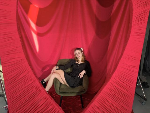 TracyLeex with blonde hair, black dress, and black heels, sitting in green chair, legs crossed, red heart-shaped curtain, smiling, hands on chair, white headband, bright lighting.