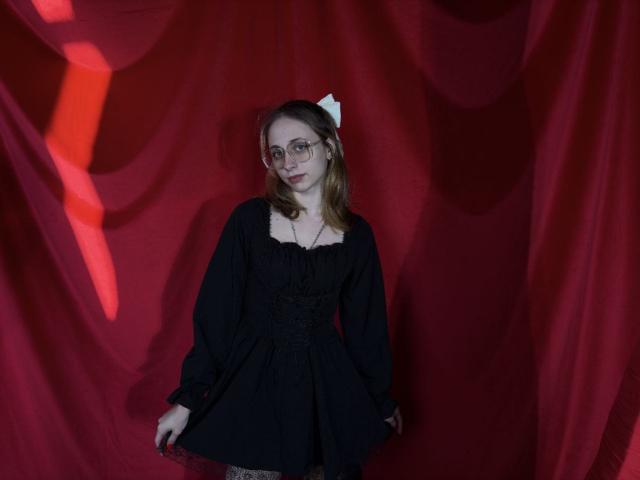TracyLeex with glasses, wearing a black dress, white bow in hair, standing against red fabric, holding dress's hem, looking slightly to the side, medium-length brown hair.