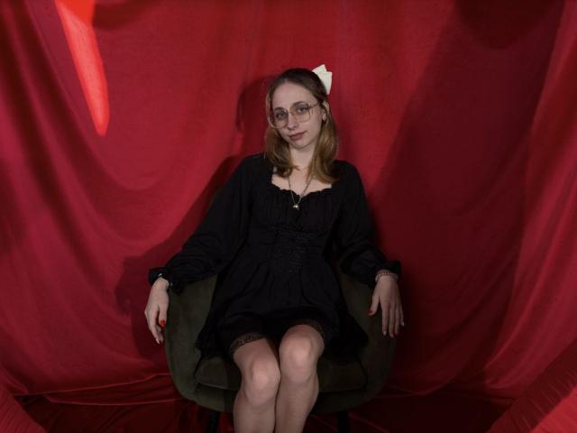 TracyLeex with glasses, black dress, and white bow, sitting on green chair, red backdrop, legs crossed, arms resting on chair, smiling, red nails, fair skin, brown hair.