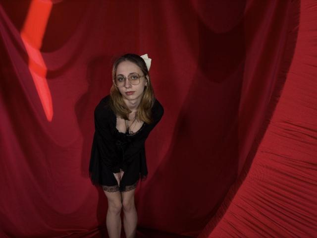 TracyLeex with glasses, black dress, and white bow, leaning forward, showing her pussy, standing against red fabric, smiling.