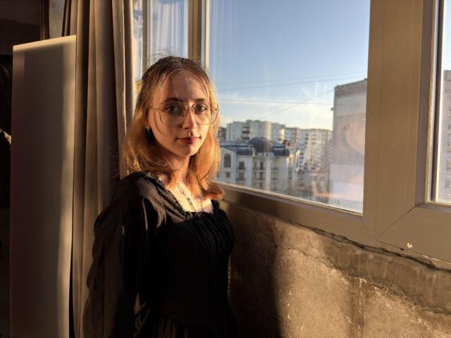 TracyLeex with glasses, light brown hair, black dress, standing by sunlit window, showing tits in shadow, city buildings visible outside, golden hour, textured wall, serious expression.