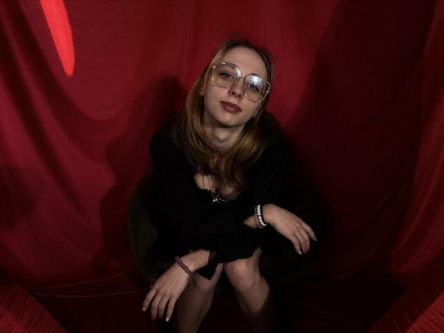 TracyLeex with glasses, black dress, and bracelet, sitting against red fabric, looking down with a slight smile, showing her ass and tits, in a dimly lit room.
