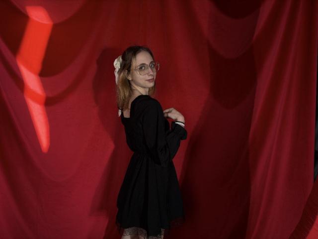 TracyLeex stands in a dimly lit setting, facing slightly away from the camera with a gentle smile. She wears a black long-sleeve dress and has a white flower in her wavy brown hair. Her glasses have small, decorative patterns. Red, draped fabric fills the background, with a bright red light casting a shadow on her right side. Her right hand is raised, fingers slightly curled. The image has a soft, shadowy quality.