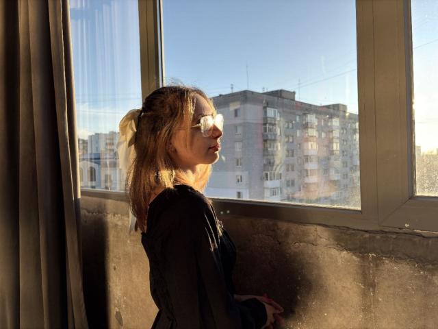 TracyLeex with blonde ponytail, wearing glasses and black top, stands by sunlit window, casting shadow on wall, looking outside at urban apartment building.