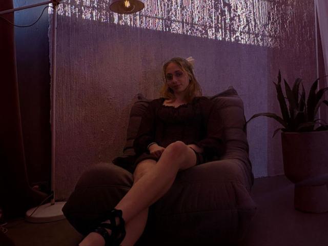TracyLeex sitting in a gray chair, wearing a dark dress, black heels, and a smile, with legs crossed, in dim purple light, looking relaxed, and with blonde hair.
