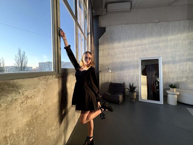 TracyLeex stands near a large window, right arm raised. She's wearing a black dress and holding a black bag in her left hand. Her blonde hair is loose, and she's wearing black ankle boots. Sunlight casts shadows on the concrete wall. A full-length mirror on the right reflects part of the room. A brown chair, potted plant, and white wall are also visible. The room has a minimalistic, industrial feel. The light and shadows create a strong contrast. TracyLeex's pose is confident and relaxed.