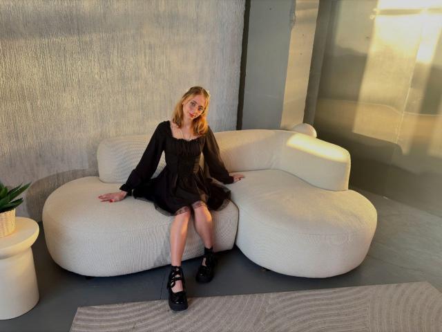 TracyLeex, blonde, sitting on white, curvy sofa, wearing black dress, off-shoulder, long sleeves, black high heels, legs spread, hands on sofa, pouty face, soft light, gray room.