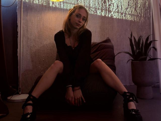 TracyLeex sits on a dark brown chair with legs spread apart. She has blonde hair, wears glasses, and a black long-sleeve top. Her black shoes have criss-cross straps. Her hands rest on her thighs, and she has red nail polish. A textured white curtain is in the background, and a potted plant is on the right. The lighting is dim, creating shadows.