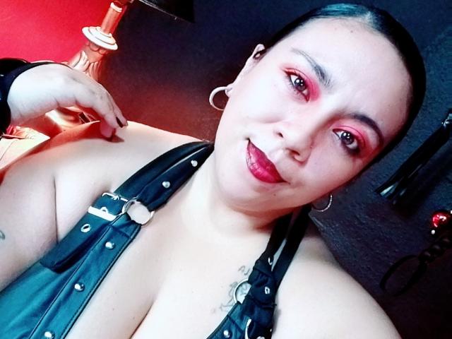 LexyDom ha l'ombretto rosso, i capelli neri, orecchini a cerchio e una giacca di pelle nera con borchie metalliche, che lascia intravedere il seno e ha un tatuaggio vicino al décolleté. Sta sorridendo..