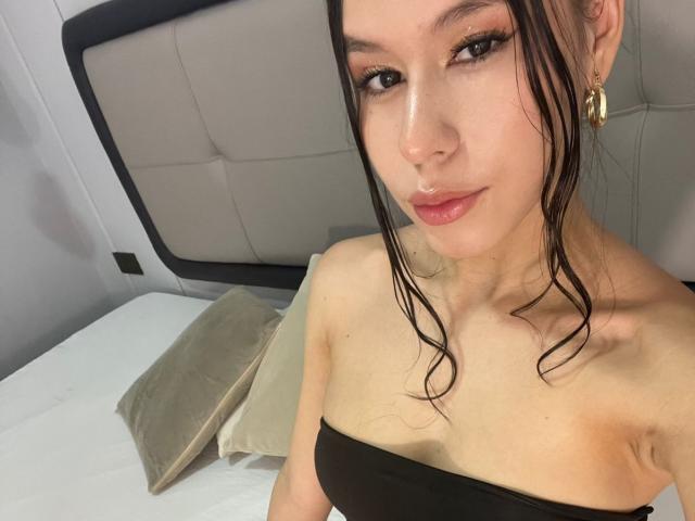 AmbarStoesselmml with wet brown hair, wearing a black strapless top, showing small tits, light skin, and a subtle nose ring, sitting on a bed with beige pillows.