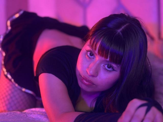 RavenLuxx with black shirt, short skirt, and fishnet stockings, on bed, butt up, looking at camera, purple and pink neon light, black nails, purple eyes, brown hair.