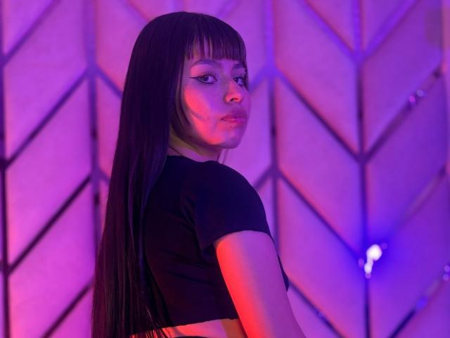 RavenLuxx with long black hair, wearing a black crop top, looking over her shoulder, purple and blue neon lights on her face, standing in front of a geometric wall.
