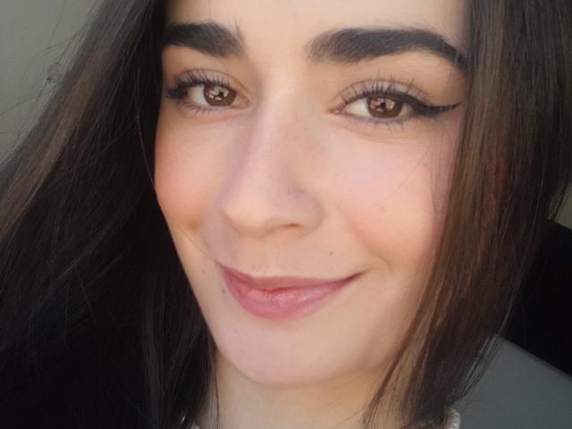 EllieBonneth's face with brown eyes, dark eyebrows, long eyelashes, pink lips, and brown hair, smiling slightly. She's got a cute nose and a small beauty mark on her chin.