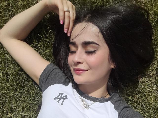 EllieBonneth with dark hair lying on grass, eyes closed, slight smile, hand touching head, wearing gray and white raglan shirt, pink nails, necklace, sunlit, relaxed.