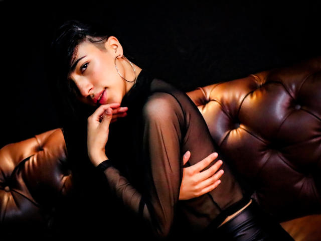 ViolettBianchi with dark hair, wearing a sheer black top, large hoop earrings, and black leather pants, lying on a brown tufted leather couch, touching her red lips.