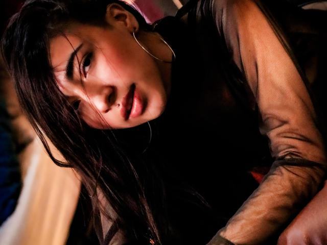 ViolettBianchi with dark, wet hair, hoop earrings, and a sheer black top, looking seductively at the camera, her small nose pierced, lips slightly parted, in a dimly lit room.