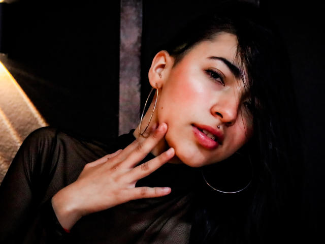 ViolettBianchi with dark hair, pink lips, nose ring, and hoop earrings, wearing a sheer black top, touching her cheek with her hand, looking seductive.