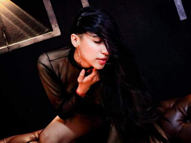 ViolettBianchi with long black hair, wearing a sheer black top, touching her chin, nose ring, and hoop earrings, sitting on a brown leather couch, dark background.