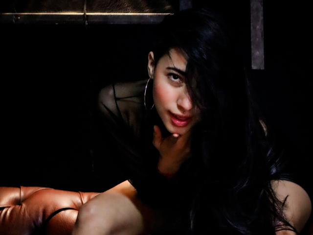 ViolettBianchi with long black hair, hoop earrings, and dark sheer top, leaning on orange leather, sultry expression, right hand on chin, soft lighting, dark background.