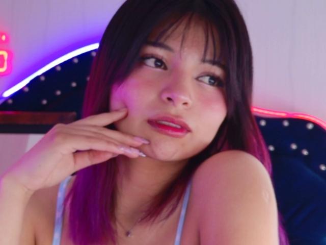 ShofiaWett with pink-tipped dark brown hair, light pink lipstick, and a cute expression, touching her cheek with one hand, wearing a light blue tank top, looking slightly to the side.
