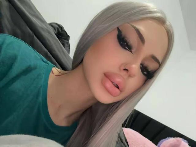 ValerieIce with long silver hair, heavy black eyeliner, and pink lipstick, wearing a green shirt, looking at the camera with slightly parted pink lips.