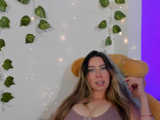 JennyLawry, con lunghi capelli castani, occhi verdi e un seno di media grandezza, indossa una maglia viola, è seduta appoggiata a un orsacchiotto giallo, tiene i capelli tra le mani, con foglie verdi e lucine..