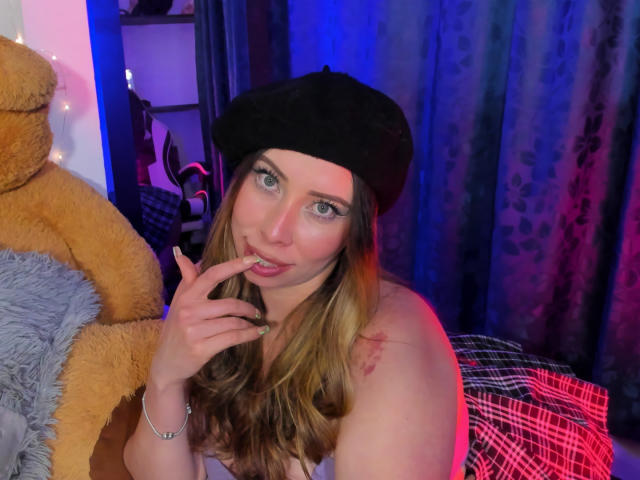JennyLawry with long brown hair, black beret, and finger in mouth, showing tattoos on her shoulder, next to a giant teddy bear, in a colorful, blue-lit room.