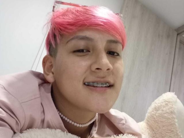 DannyRouds with pink hair, wearing a pink shirt and pearl necklace, has metal teeth and is holding a stuffed teddy bear, smiling.