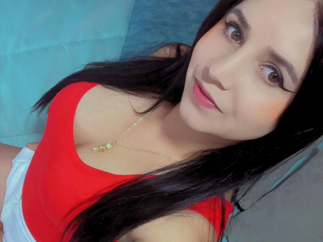 Kkatalina with long black hair, brown eyes, and red top showing her tits, wearing a gold necklace, smiling slightly. She's sitting on a blue surface.