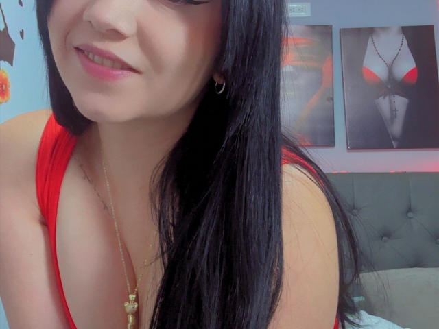 Kkatalina with long black hair, red dress, gold necklace, smiling, showing cleavage, small hoop earring, red lipstick, soft light, bedroom background with art.