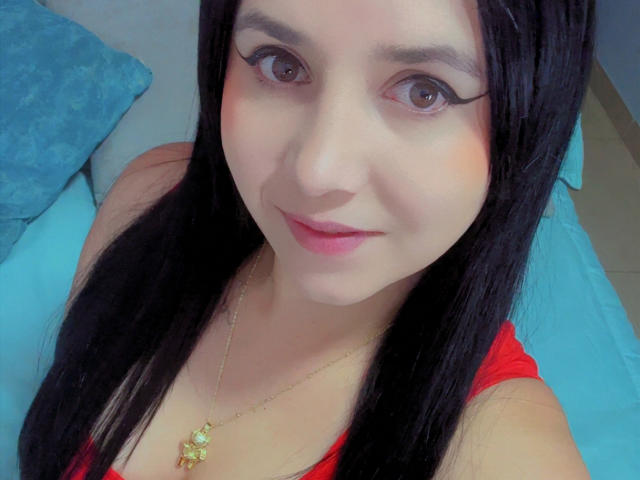 Kkatalina has long black hair, brown eyes with thick eyeliner, red lipstick, and a gold necklace with a flower pendant. She's wearing a red top, smiling at the camera.