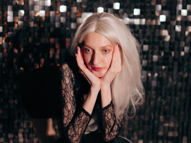 ViktoriaClark with platinum blonde hair, black lace top, hands on face, leaning forward, looking into camera, subtle smile, shiny sequined background, soft lighting, confident, sexy, cute.