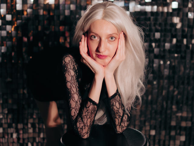 ViktoriaClark with long platinum blonde hair, hands on her face, wearing a black lace top, looking at the camera with blue eyes, sitting against a shiny black background.