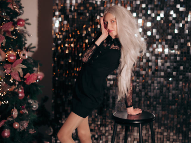 ViktoriaClark with long white hair, black dress, and lace sleeves, leaning on a stool, hand on cheek, next to a Christmas tree with pink ornaments.