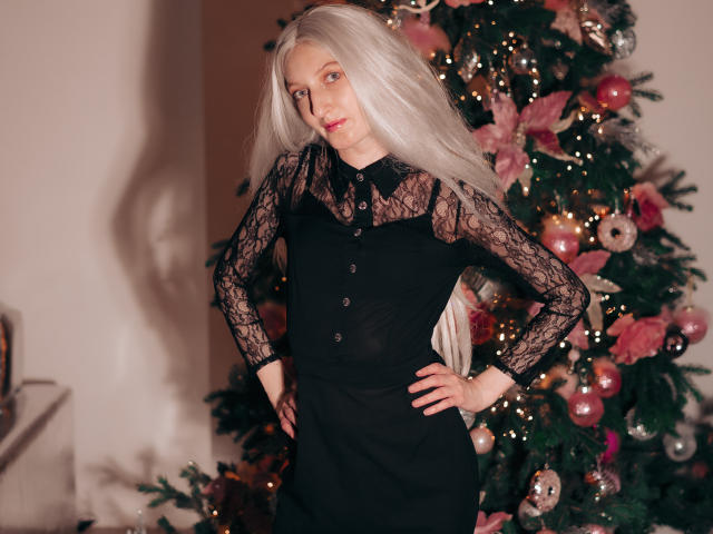 ViktoriaClark with long white hair, black lace long-sleeve dress, hands on hips, standing in front of a decorated Christmas tree, looking slightly to the side, smiling subtly.