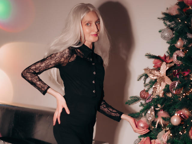 ViktoriaClark with long white hair, black lace-sleeved dress, standing by decorated Christmas tree, hand on hip, looking at camera, wearing black buttons on dress, smiling subtly.