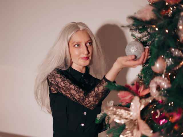 ViktoriaClark with long white hair, black lace top, and pink lipstick, holding a silver ornament, decorating a Christmas tree with pink and gold ornaments.