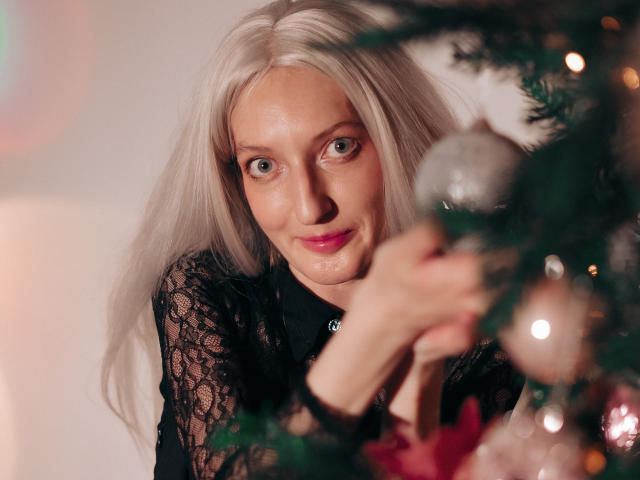 ViktoriaClark with long blonde hair, black lace top, and pink lipstick, holding a Christmas ornament, smiling softly. Boob outline visible through lace. Christmas tree in foreground.