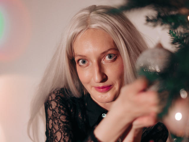 ViktoriaClark with blonde hair, blue eyes, and pink lipstick, wearing a black lace top, gently holding a Christmas ornament, smiling slightly. Her boobs are not visible.