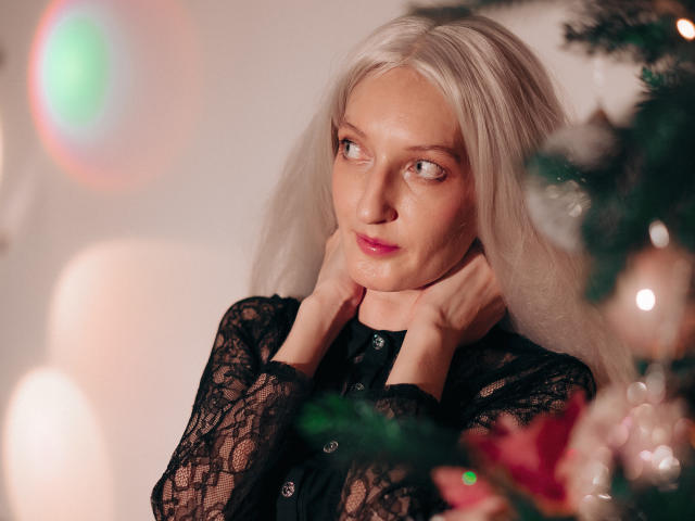 ViktoriaClark with long, white hair, wearing black lace, hands on neck, looking to the side, Christmas tree in foreground, soft light, pink lips, blue eyes, slightly pouty expression.