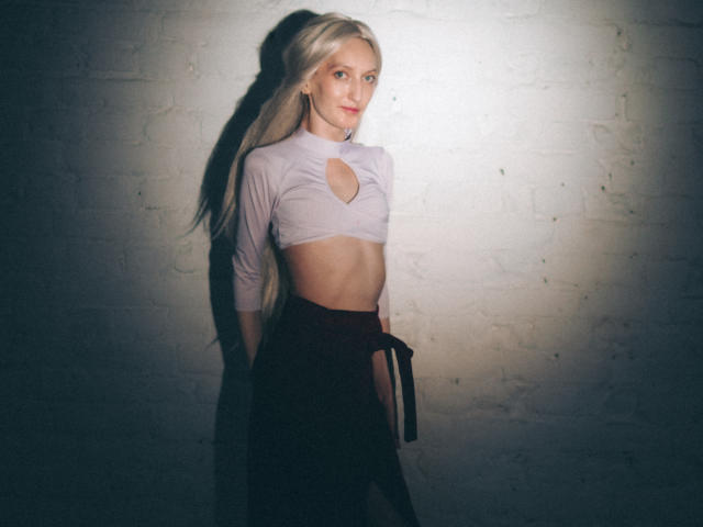 ViktoriaClark has blonde hair, a slim build, and small tits. She's wearing a cut-out, long-sleeve top and a dark skirt, standing against a white brick wall.