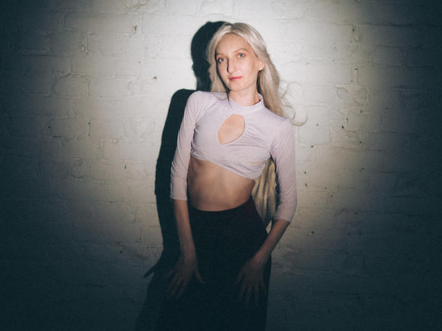 ViktoriaClark with long blonde hair, wearing a white, long-sleeve, cut-out top, exposing her small tits, standing against a white brick wall, looking at the camera.